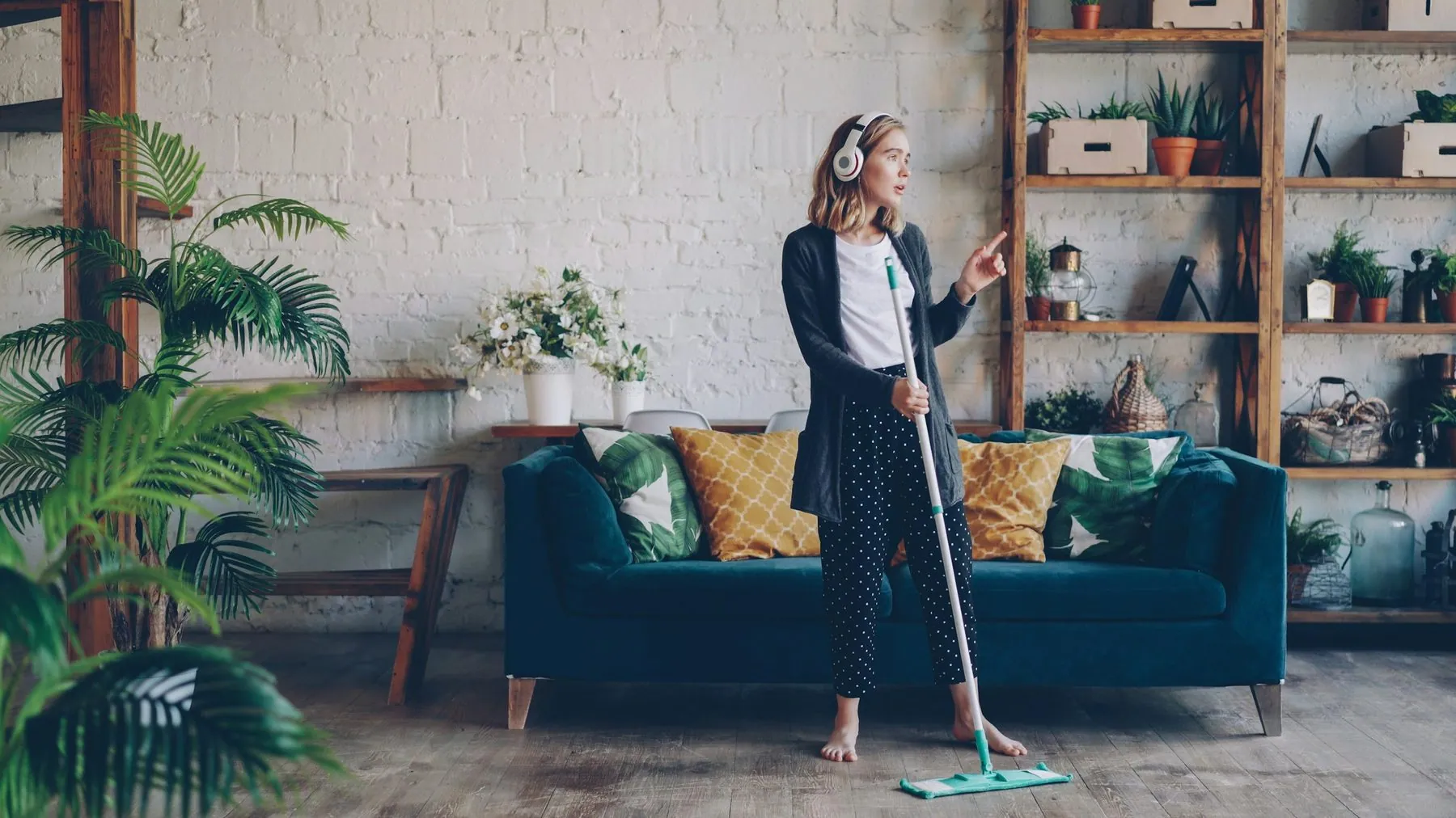 Beautiful young lady is listening to music through wireless headphones and doing housework cleaning the floor with mop.