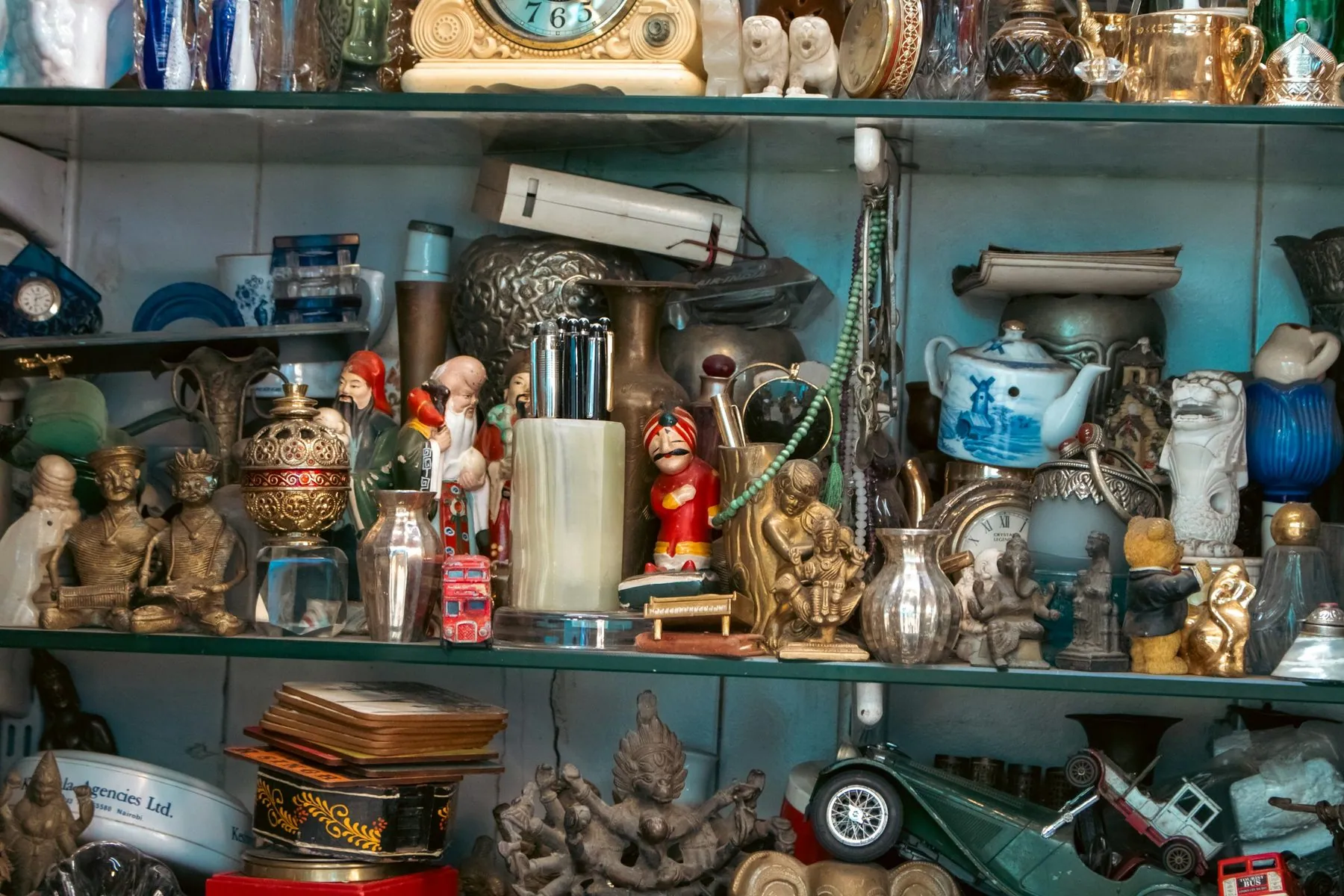 A detailed photograph of a crowded antique shop shelf filled with figurines, brass statues, teapots, clocks, vases, toys