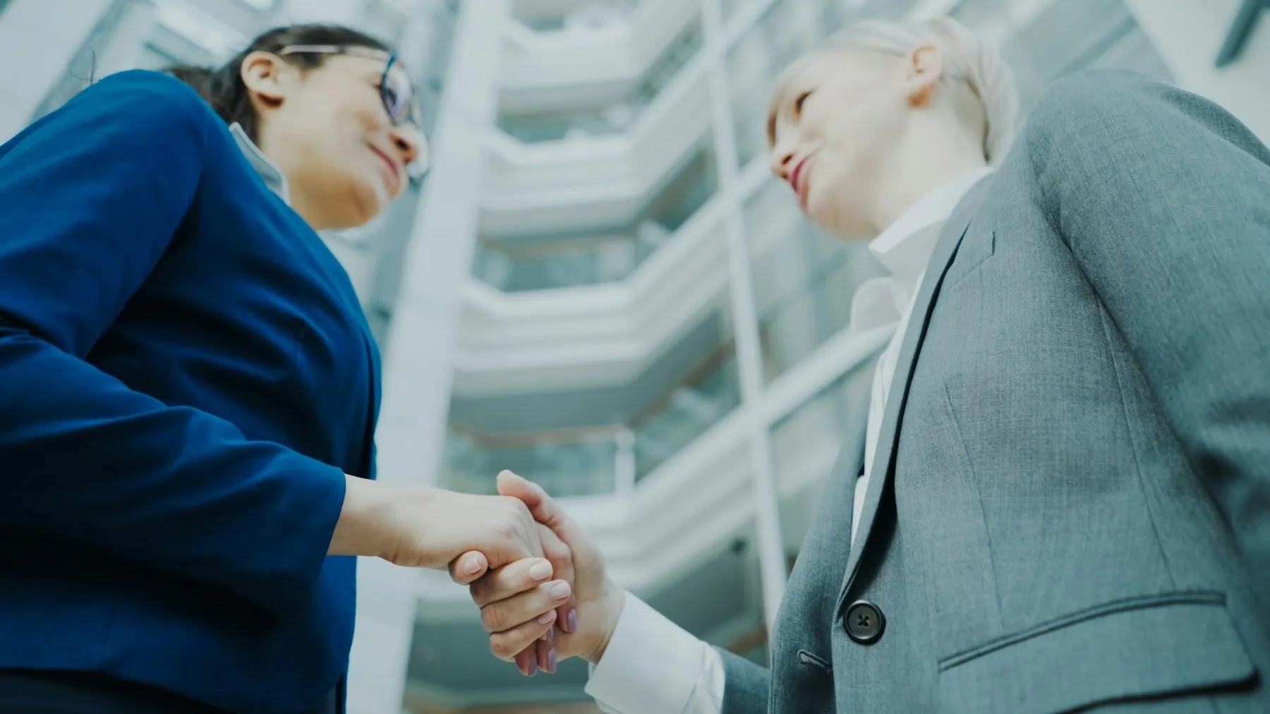 Low angle shot of businesswoman shaking hands and talking with female business colleague in suit in hall of modern offic