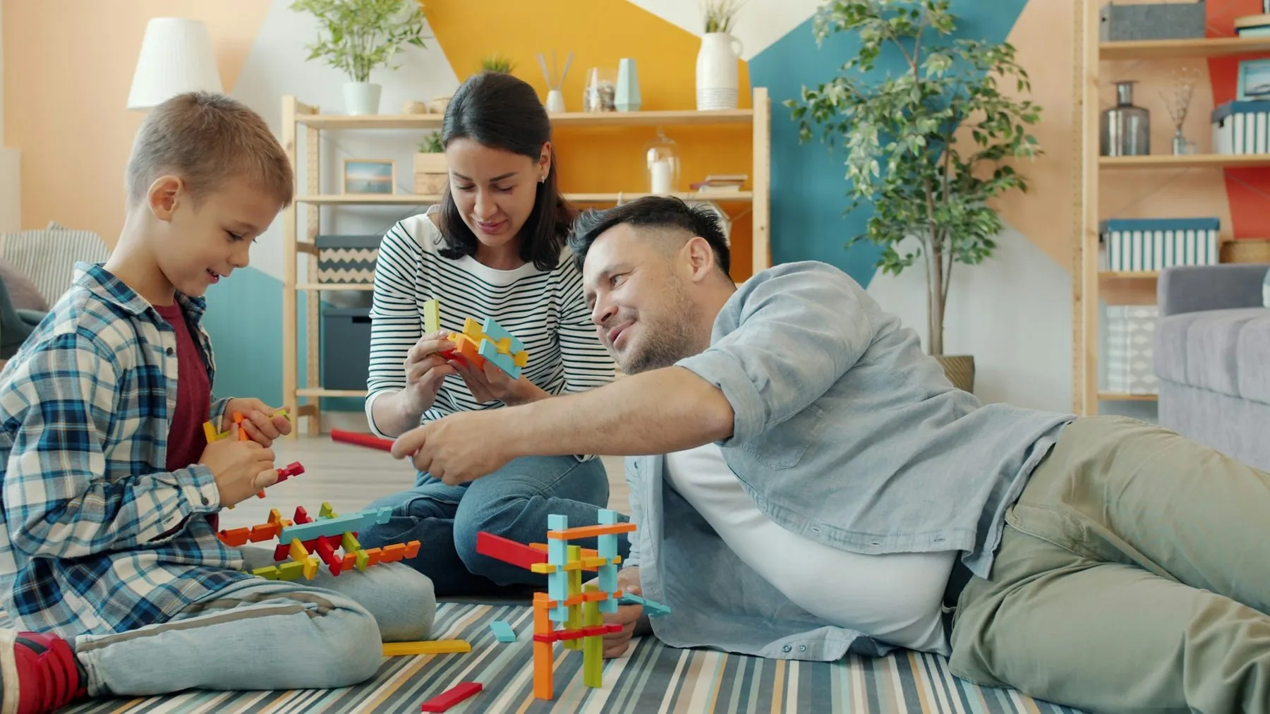 Child is playing with mother and father cheerful adults at home building with construction blocks sitting on floor toget