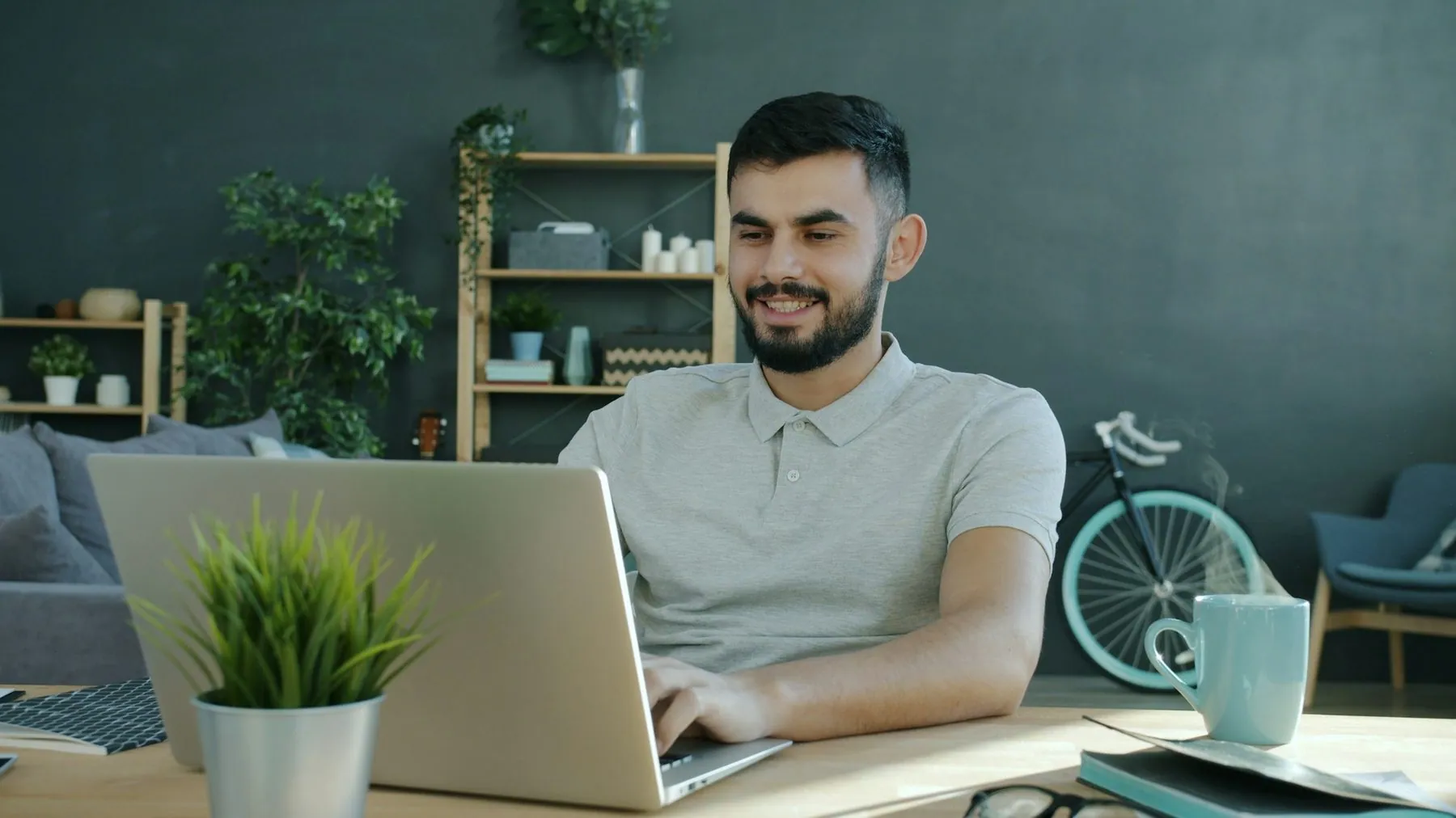 Cinemagraph of Middle Eastern man freelancer working with laptop and smiling while steam rising from hot cup in apartmen