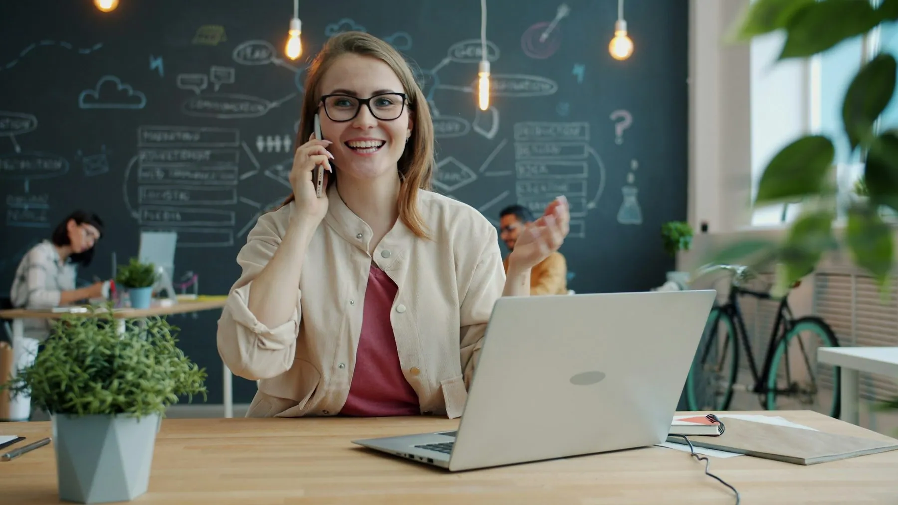 Young business woman attractive blonde is talking on mobile phone in shared office discussing project sitting at desk wi