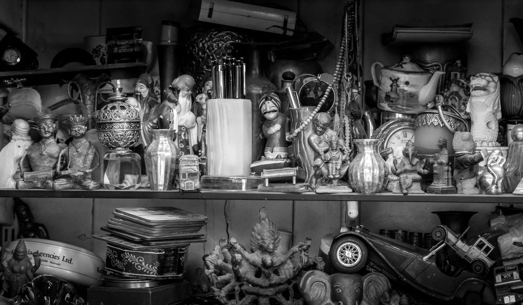 A detailed photograph of a crowded antique shop shelf filled with figurines, brass statues, teapots, clocks, vases, toys