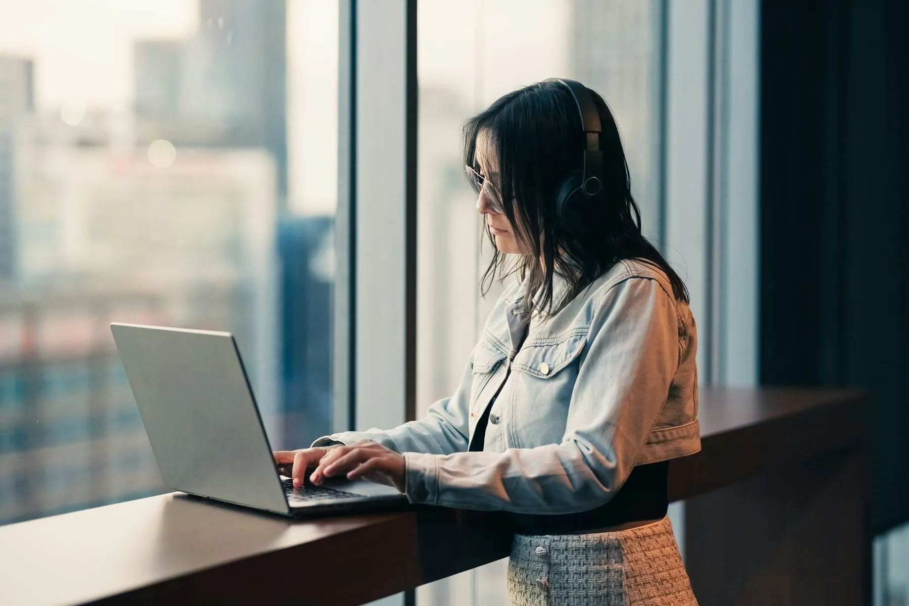 Woman wearing headphones works on a laptop by window