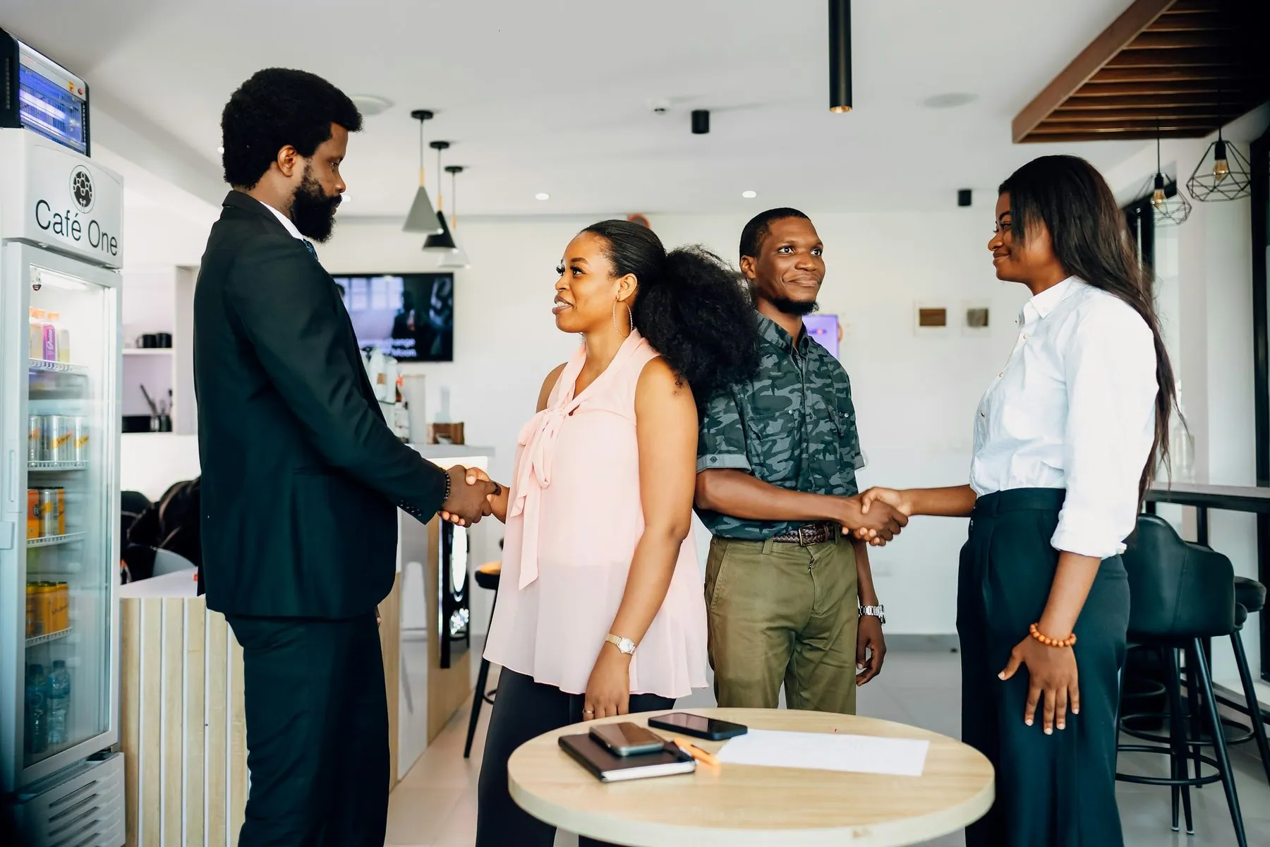 A group of four business people are standing around a table shaking hands and talking.