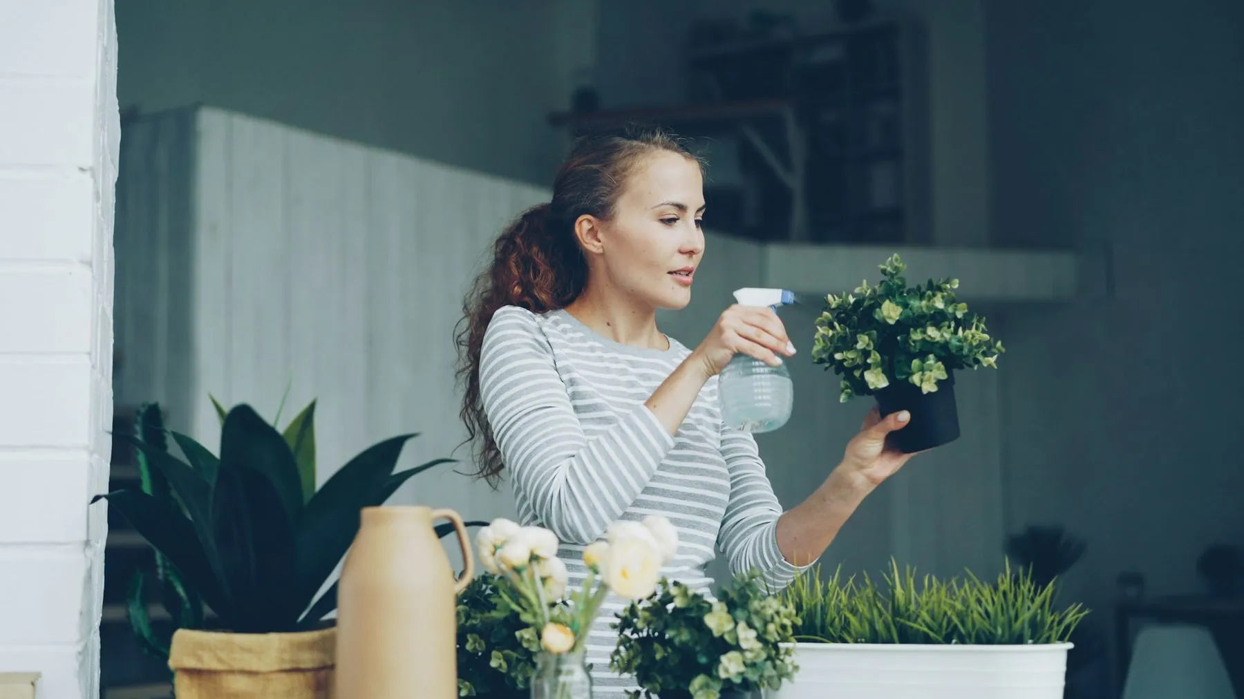 Cheerful housewife is watering green plants using sprayer holding flowerpots and smiling standing in beautiful light apa