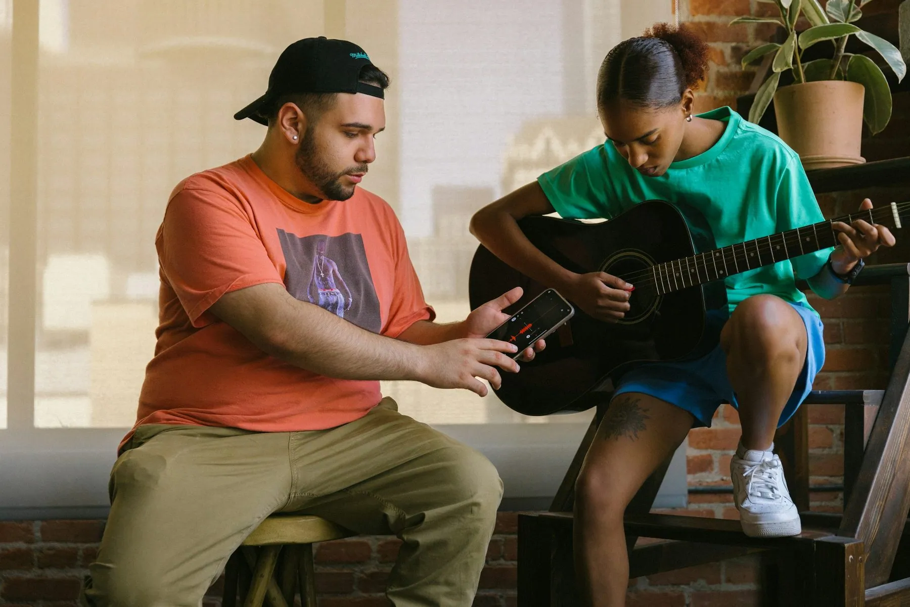 man in red shirt and woman in green shirt recording music on phone and playing the guitar