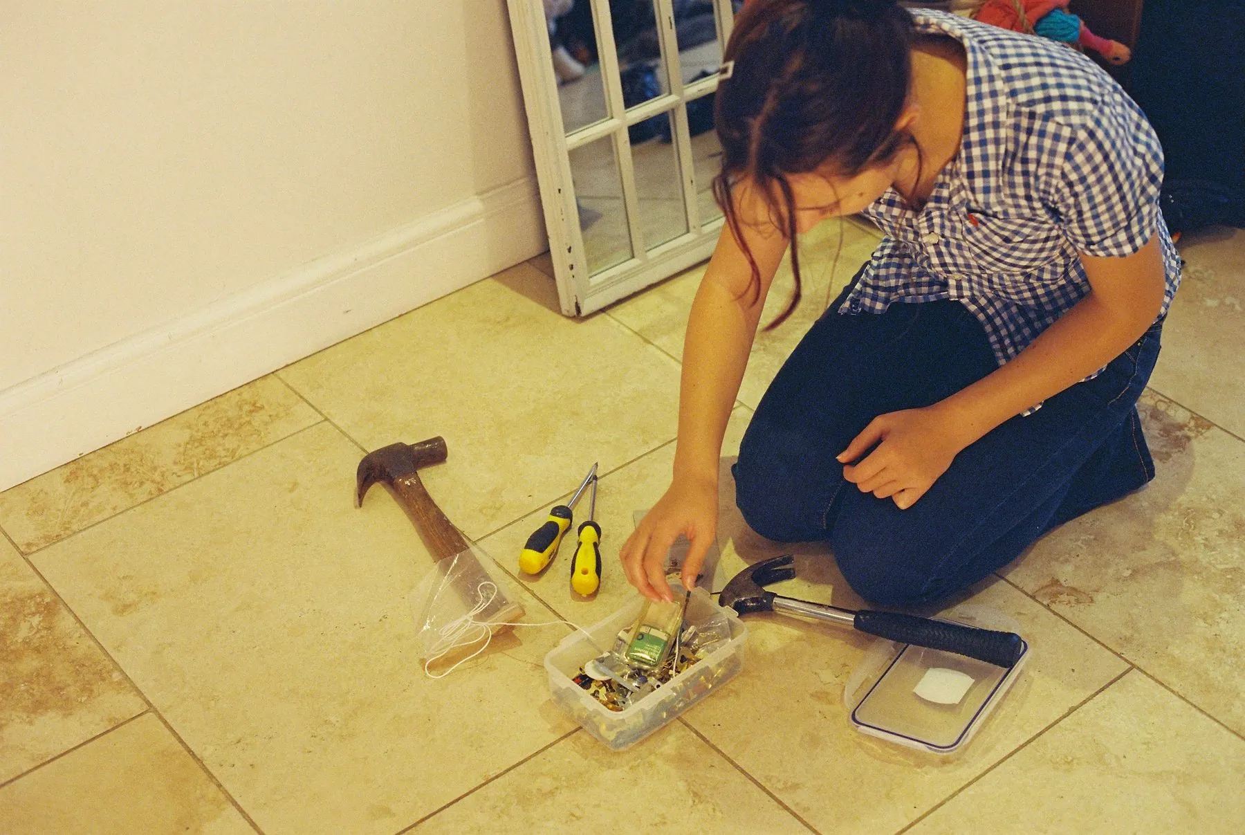 Kneeling on a tiled floor, a woman sorts through a box of fixings with a hammer and screwdrivers beside her, ready to ha