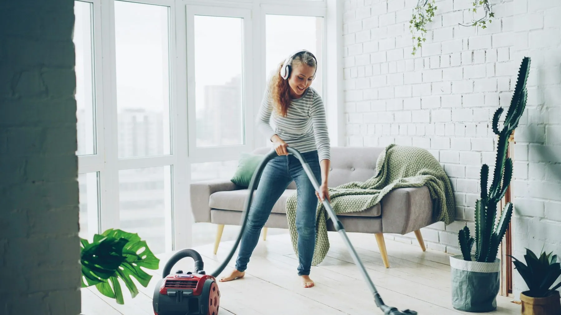 Attractive young lady is dancing and singing listening to music through wireless headphones during housecleaning at home